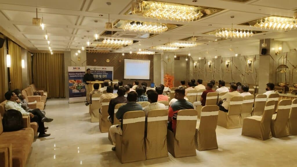 A conference room with attendees seated and a speaker presenting at the front, under elegant chandeliers.