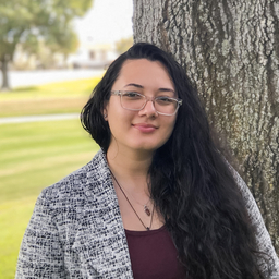 A woman wearing glasses and a blazer leaning against a tree.