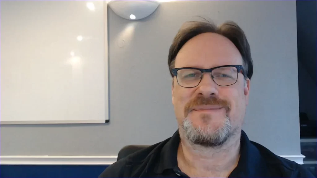 A man in glasses is smiling in front of a white board.